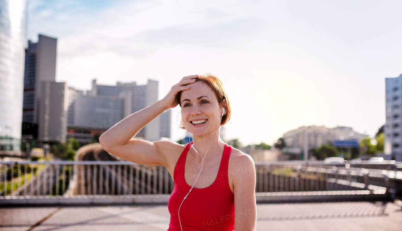 Front view of young woman runner with earphones in city, resting on the bridge.