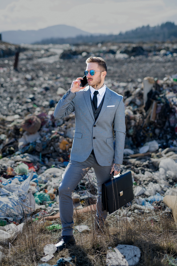 Fashionable modern businessman with smartphone on landfill, consumerism versus pollution concept.