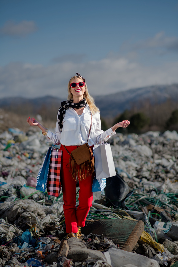 Fashionable modern woman with shopping bags on landfill, consumerism versus pollution concept.