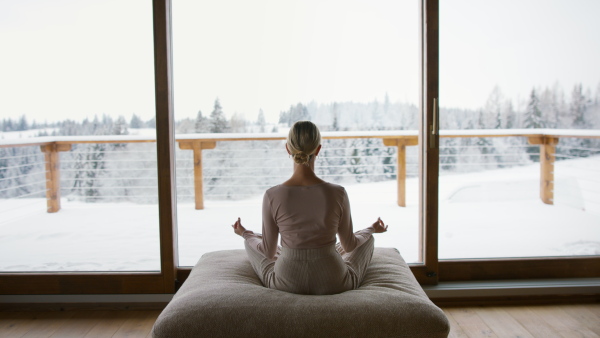 A rear view of woman sitting and exercising yoga indoors in winter apartment.