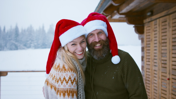 A happy couple in santa hat kissing and looking at camera outdoors in winter nature, holiday at Christmas time.