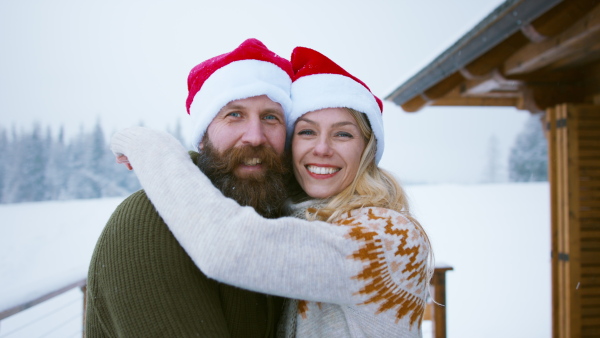 A happy couple in santa hat kissing and looking at camera outdoors in winter nature, holiday at Christmas time.