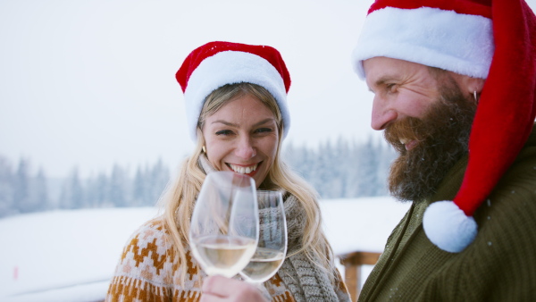 A couple in santa hats clinking wine glasses and talking outdoors in winter nature at Christmas time