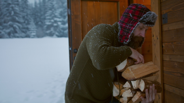 A mature man holding firewood outdoors in front of cottage in winter time.