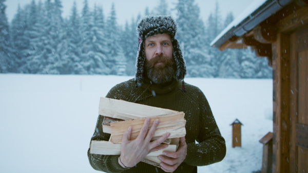 A mature man holding firewood outdoors in front of cottage in winter time, looking at camera.