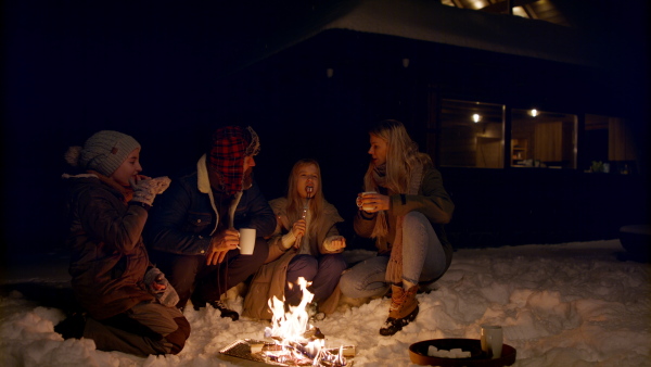 A family with two small daughters roasting marshmallows on campfire outdoors by cottage in winter at night.