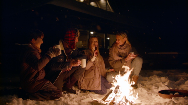 A family with two small daughters roasting marshmallows on campfire outdoors by cottage in winter at night.