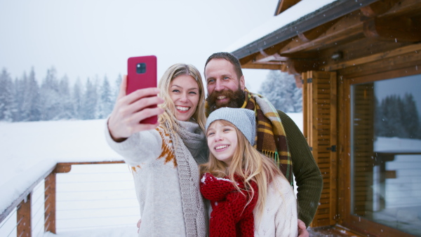 A happy family with small daughter taking selfie and making faces by mountain hut outdoors in winter.