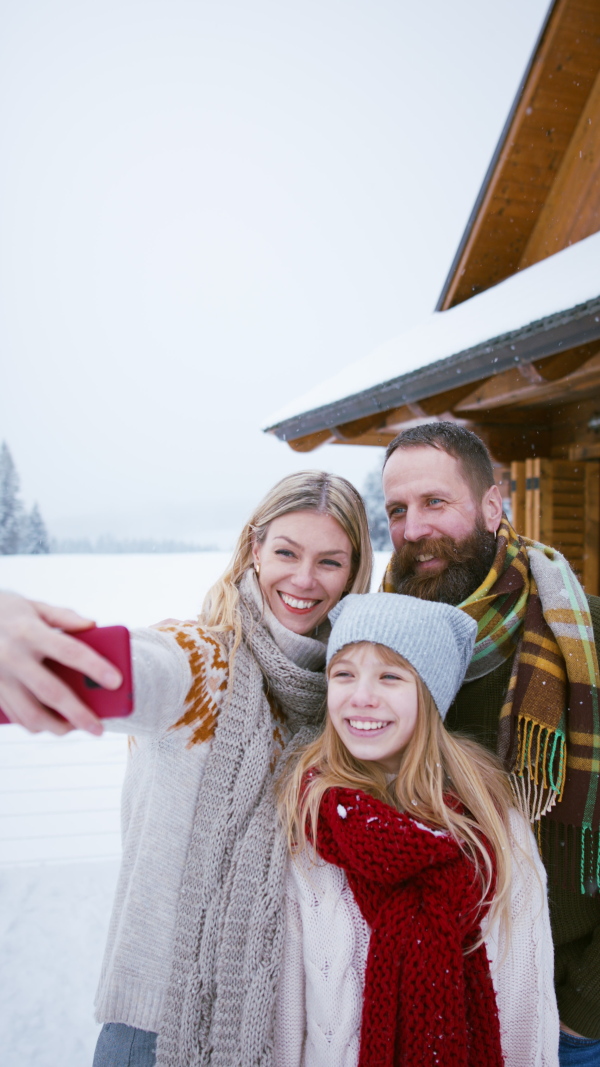 A vertical footage of happy family with small daughter taking selfie and making faces by mountain hut outdoors in winter.