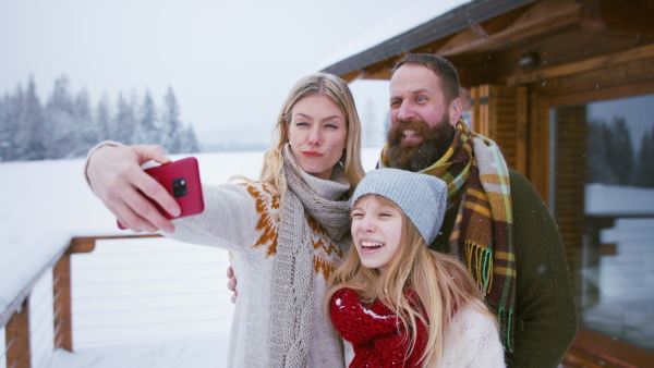 A happy family with small daughter taking selfie and making faces by mountain hut outdoors in winter.