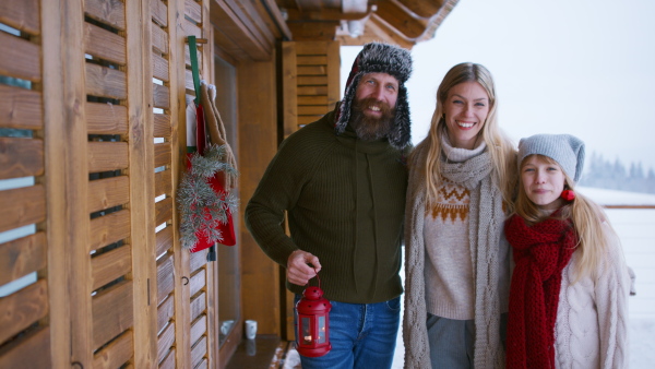 A happy family with small daughter looking at camera by mountain hut outdoors in winter.