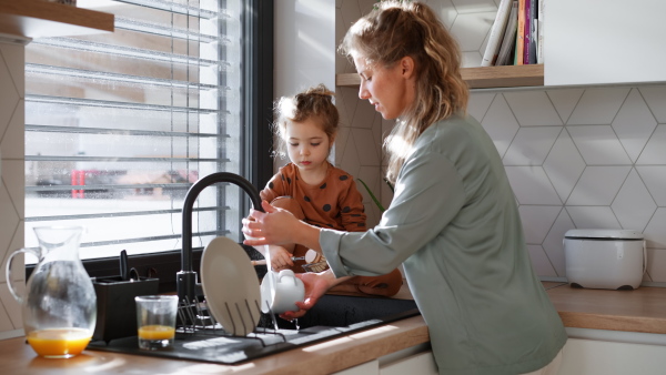 A young mother with little daughter washing dishes together in kitchen