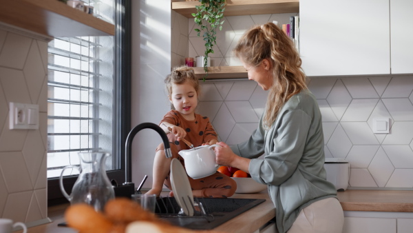 A young mother with little daughter washing dishes together in kitchen