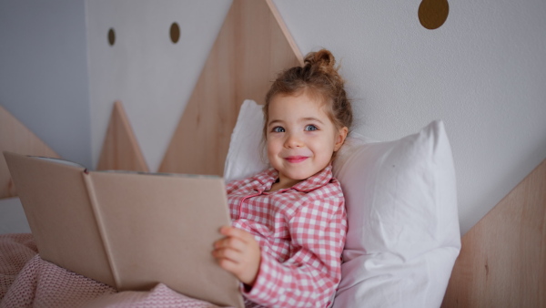 A cute happy litttle girl in pyjamas sitting in bed and reading book.