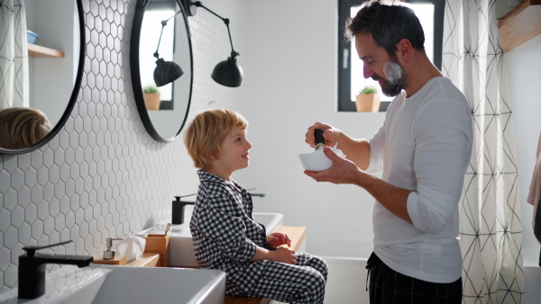 A father with small child indoors in bathroom in the morning at home, shaving.