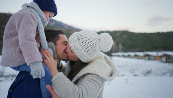 A happy young family on winter holiday with small child kissing outdoors in snowy nature.