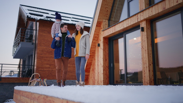 A low angle view of happy young family on holiday with small child standing in front of cabin in winter.