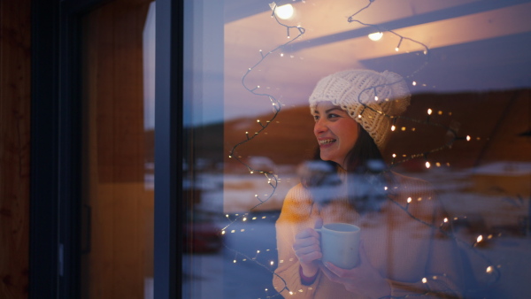 A happy young woman with tea looking throug window, winter holiday in cabina.