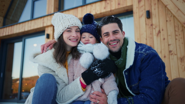 A happy young family on winter holiday in cabin with small child outdoors in snowy nature, looking at camera.