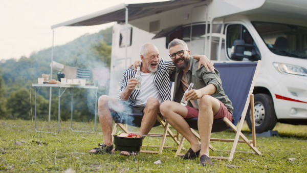 A mature man with senior father talking at campsite outdoors, barbecue on caravan holiday trip.