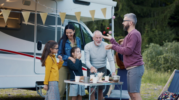 A multi-generation family eating together outdoors by car, caravan holiday trip. Man joggling with tomatoes.