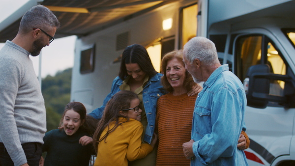 A multi-generation family looking at camera outdoors at dusk, caravan holiday trip.