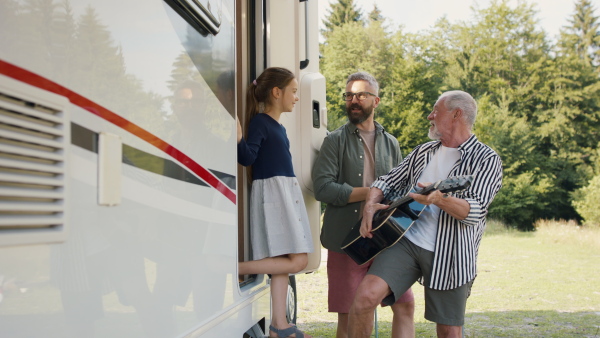 Happy multi-generation family unpacking and grandfather playing on guitar by car, caravan holiday trip.