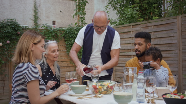 A multiracial three generations family sitting and eating lunch together outdoors in back yard.