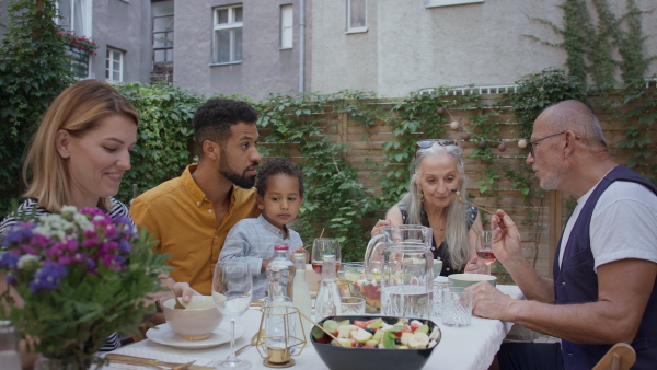 A multiracial three generations family sitting and eating lunch together outdoors in back yard.