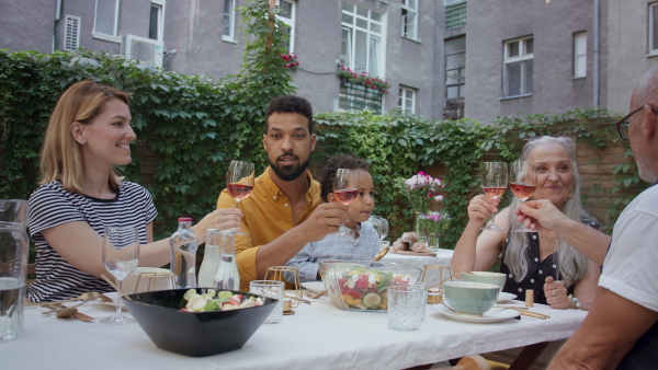 A multiracial three generations family sitting and clinking glassesbefore lunch outdoors in back yard.