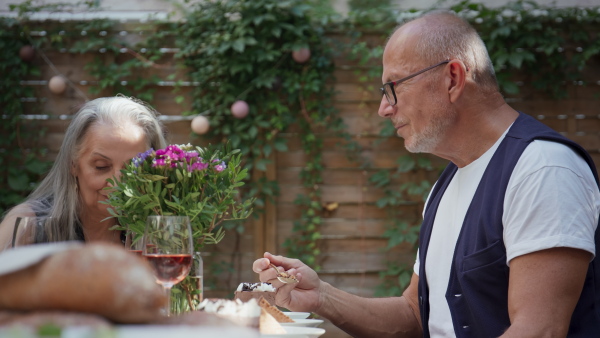 A senior couple sitting and eating cake during birthday party outdoors in back yard.