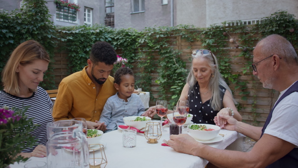 A multiracial three generations family sitting, holding hands and praying together before lunch outdoors in back yard.