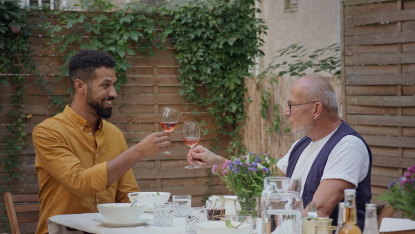 A young African American man with his father in law sitting and clinking wine glasses outdoors in back yard.