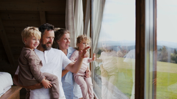 A happy young family in pajamas with small children looking at view from window on holiday.