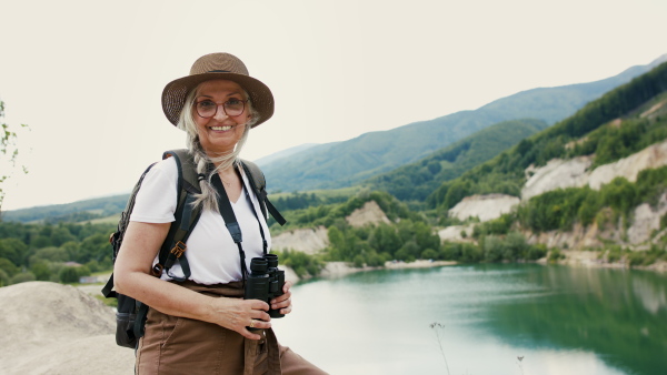 A happy senior woman tourist on hiking trip on summer holiday, standing by lake and looking through binoculars