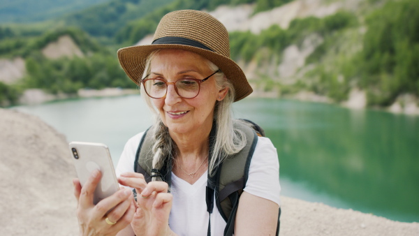 A happy senior woman in sun hat with smartphone hiking in nature by lake.