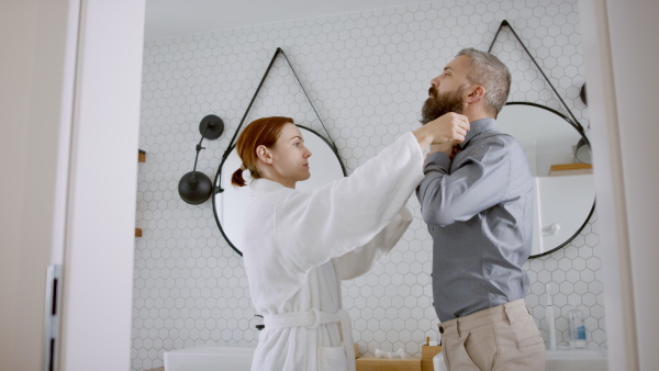 Happy couple in bathroom getting ready for work in the morning. Wife helping husband with shirt.