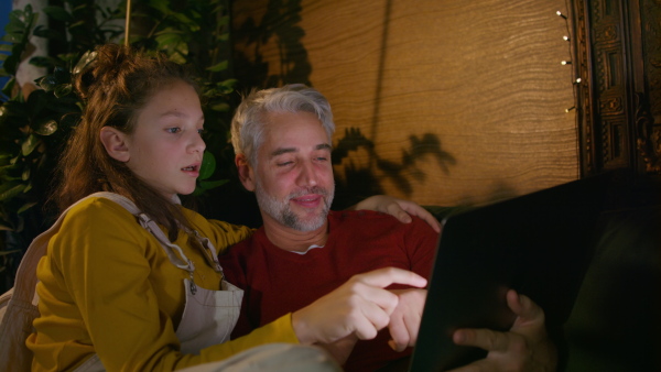 A father sitting on sofa with daughter and using laptop together at home in evening.