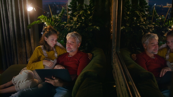 A father sitting on sofa with daughter and using laptop together at home in evening.