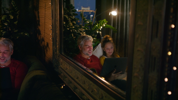 A father sitting on sofa with daughter and using laptop together at home in evening.