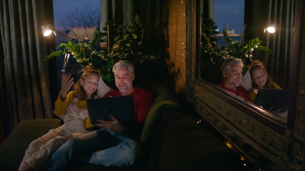 A father sitting on sofa with daughter and using laptop together at home in evening.