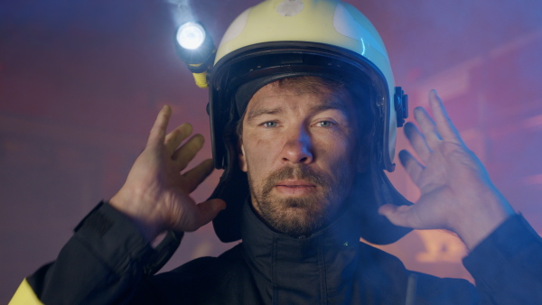 A close up of mature dirty firefighter putting on helmet with fire truck at background at night.