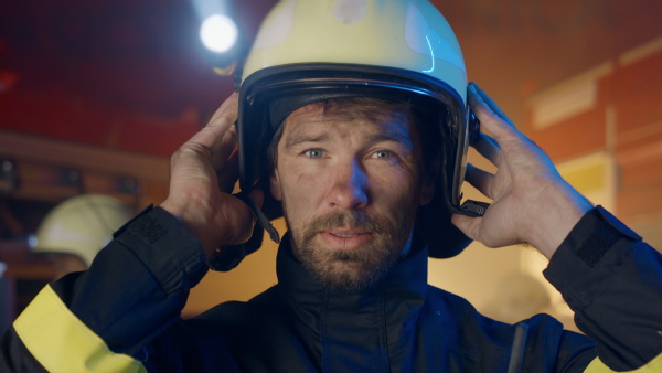 A close up of mature dirty firefighter putting on helmet with fire truck at background at night.