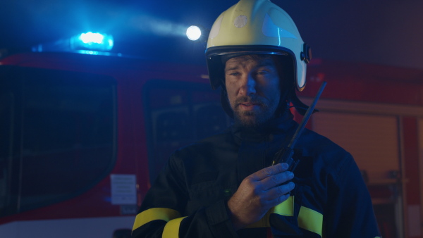 A mid adult firefighter man talking to walkie talkie with fire truck in background at night.