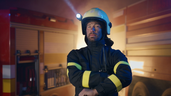 A mid adult dirty male firefighter looking at camera and smiling with fire truck at background at night.