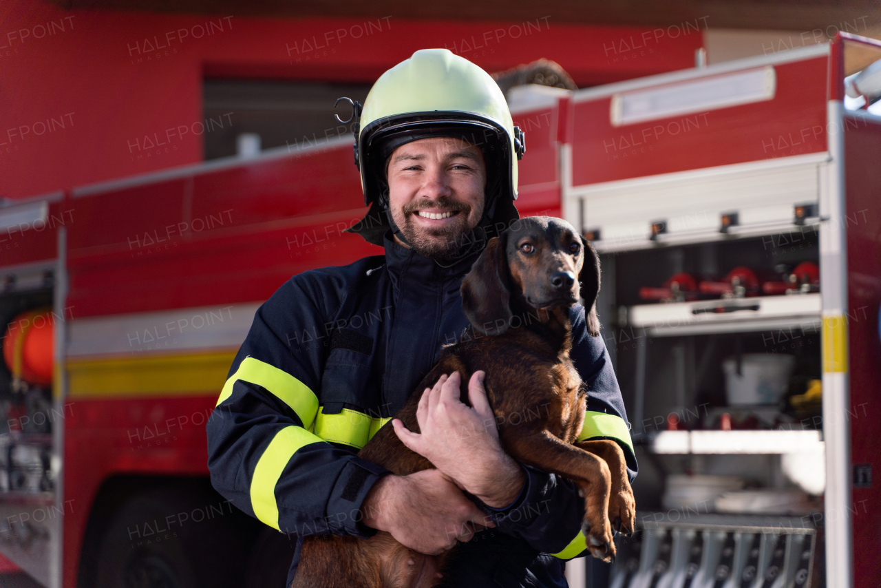 A happy mature firefighter man holding dog and looking at camera with fire truck in background