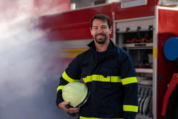 A happy mid-adult firefighter man looking at camera with fire truck and smoke in background.