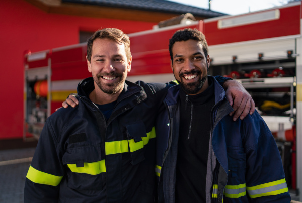 Happy firefighters crew with fire station and truck in background looking at a camera.