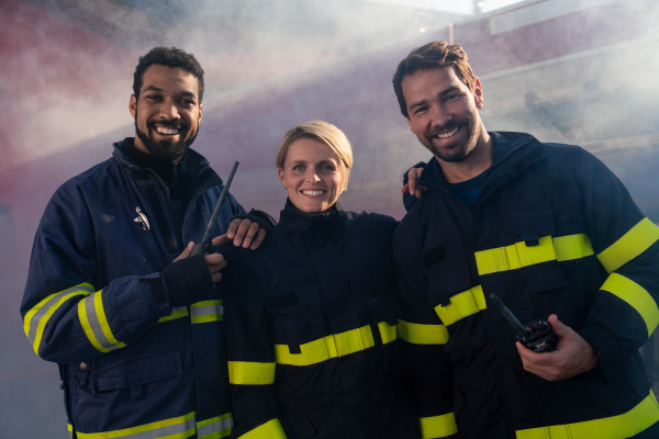 Happy firefighters crew with fire station and truck in background looking at a camera.