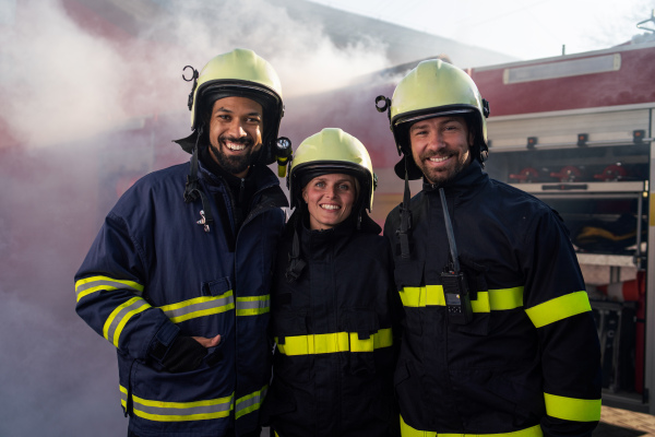 Happy firefighters crew with fire station and truck in background looking at a camera.